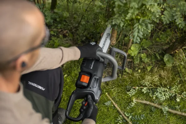 User trimming small branches with the Husqvarna Aspire C15X-P4A2 cordless chainsaw in a garden setting.