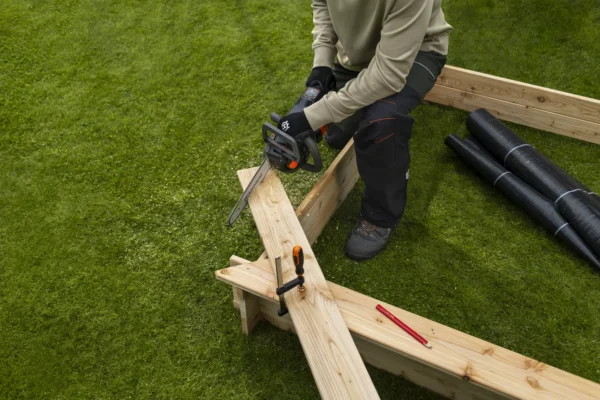 User cutting timber planks on a lawn with the Husqvarna Aspire C15X-P4A4 cordless chainsaw during a garden construction project.