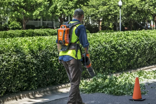 Worker using Husqvarna BLi950X Backpack Battery to power a leaf blower during hedge maintenance in a landscaped area.