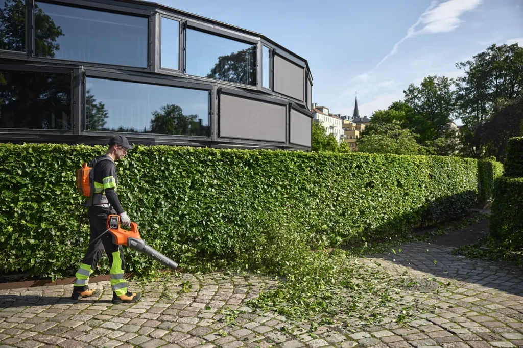 Professional landscaper using Husqvarna BLi950X Backpack Battery and leaf blower to clear trimmed hedges in an urban garden.
