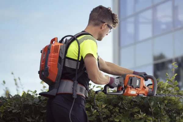 Professional landscaper using a Husqvarna BLi950X Backpack Battery with a hedge trimmer for urban shrub maintenance.